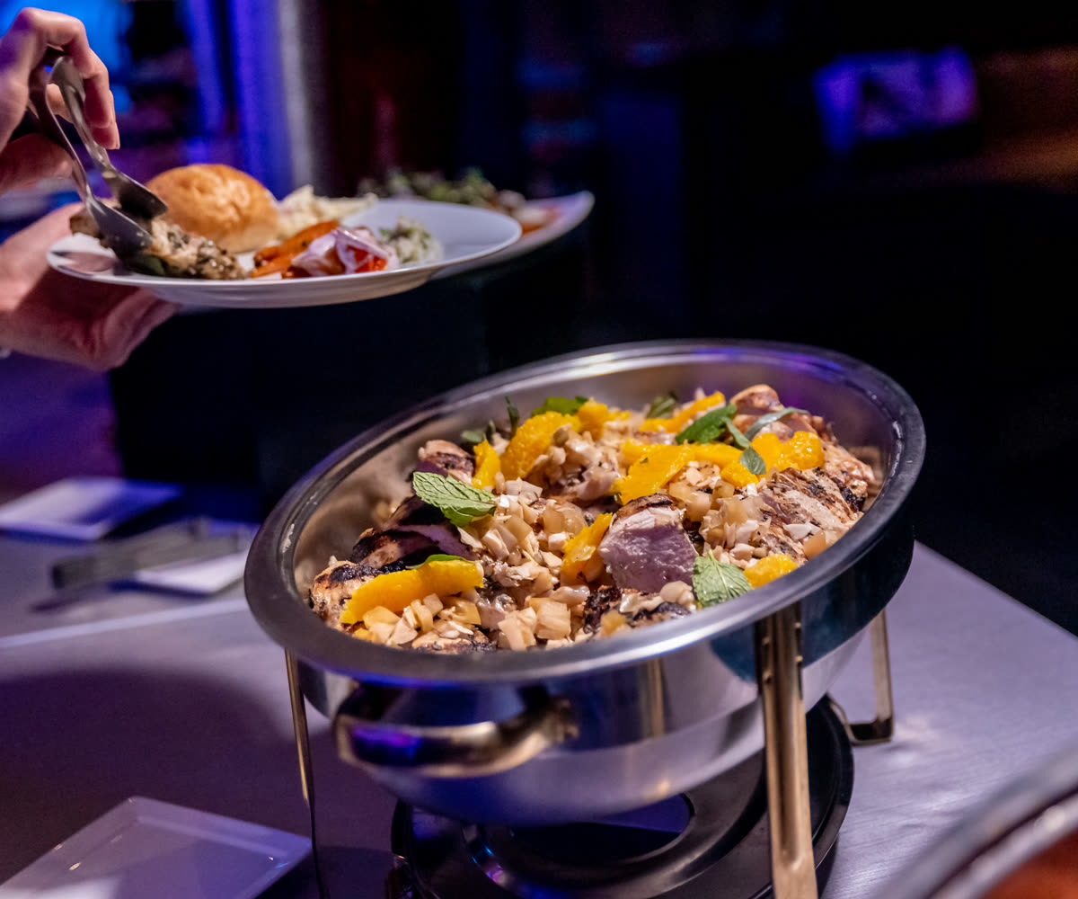 A guest serving themselves at a buffet, lifting food onto a plate beside a polished chafing dish of grilled chicken garnished with orange and mint